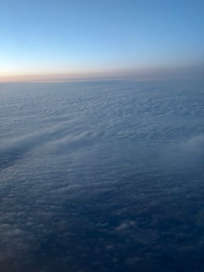 This image shows a view from high altitude, likely from an airplane or spacecraft, looking down at the Earth's atmosphere. The scene is dominated by a vast expanse of clouds, which appear textured and layered. The clouds transition from lighter, more diffuse formations on the left side of the image, where they meet a soft, hazy horizon, to darker, more densely packed formations on the right. The left side of the image also features a gradient of blue, suggesting a clear sky at a very high altitude, possibly during sunrise or sunset, indicated by a faint peachy glow along the horizon line. The cloud formations themselves are varied, with some appearing as rippled waves and others as smaller, puffy mounds. There is no visible text in the image. The overall impression is one of serene detachment from the world below, showcasing the beauty and complexity of atmospheric phenomena.