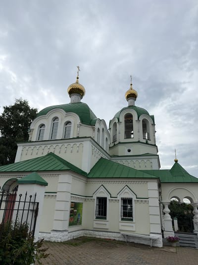 The image shows the exterior of a church in Karakulino, Russia. The church has a light-colored facade with a green roof and golden domes topped with crosses. There are arched windows and decorative architectural details. In the foreground, there is a black metal fence and some greenery. The sky is overcast.