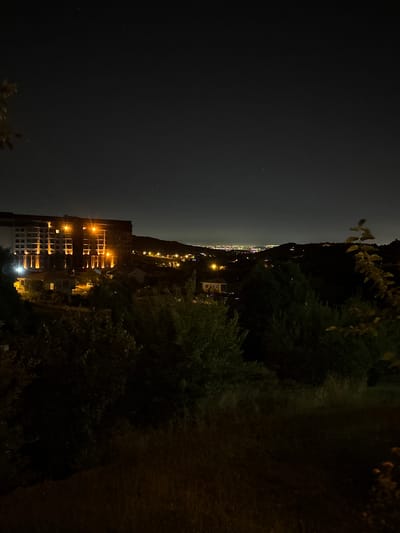 The image depicts a nighttime view of a residential area in Termal, Turkey, overlooking a distant cityscape. In the foreground, dark silhouettes of trees and vegetation obscure the lower portion of the scene. A large, multi-story building, illuminated by numerous warm orange lights, dominates the mid-ground. This building appears to be a hotel or apartment complex, with visible windows and architectural details. Smaller houses and structures are scattered around the base of the larger building, also lit by artificial lights. Beyond this immediate area, rolling hills are covered in dark vegetation, with the lights of the distant city of Termal forming a bright, horizontal band on the horizon. The sky is dark, with a few faint stars visible, suggesting a clear night. The overall impression is one of a quiet, illuminated settlement nestled within a natural landscape at night.