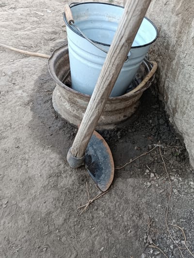 This media file captures an outdoor scene at ground level, likely in a rural or utilitarian setting. The primary objects are a light blue enamel bucket, a worn metal rim that appears to be from a wheel or a large basin, and a hoe with a dark metal blade and a wooden handle. The bucket is resting inside the metal rim, and the hoe is positioned in front of them, leaning against the rim and the bucket.
The ground is composed of dry, dusty soil mixed with small pebbles and some dried organic matter. A thin, light-colored hose or pipe is visible extending from the left side of the frame into the distance, partially obscured by the soil. The background features a rough, textured wall, possibly made of concrete or stone, which provides a backdrop to the arrangement of objects.
The scene appears to be set during the daytime, with natural light illuminating the objects. There are no people visible, and the focus is on the tools and the environment. The overall impression is one of utility and perhaps manual labor, as these are common implements for gardening or other outdoor tasks. The worn appearance of the metal rim and the hoe suggests they are well-used. The image provides no specific cues about the weather beyond what can be inferred from the dry soil. The location context of Leușeni, Moldova, adds a geographical detail but does not significantly alter the visual interpretation of the scene itself. There is no visible text in the image.