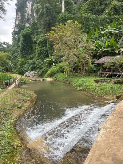 The scene depicts a rural watercourse in Ban Nampoun, Laos, situated within a valley flanked by densely vegetated limestone karsts under an overcast sky. In the foreground, a two-tiered concrete weir spans the water body, creating a cascade of aerated water flowing towards the viewer. Upstream of the weir, the water is calm and clear, with a greenish tint.
On the left bank, a dirt path runs parallel to the water, bordered by a rustic wooden railing consisting of vertical posts and horizontal rails. This path leads to a low-lying bridge upstream, constructed from three concrete pipes embedded in the bank, allowing water to flow beneath. A large, light-colored boulder rests on the bank near this pipe bridge.
On the right bank, a concrete edge or pathway is visible immediately adjacent to the weir. Further back, an elevated wooden hut or pavilion with a dark, possibly tiled roof is positioned near the water's edge. Two individuals are discernible inside the hut, appearing to be seated or reclining. The surrounding environment is characterized by dense tropical foliage, including large banana plants, various trees, and lush undergrowth covering the banks and the steep slopes leading up to the towering limestone cliffs in the background.