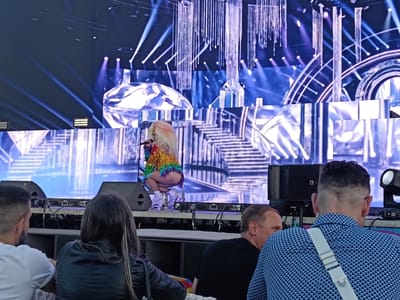 Captured at a venue, possibly in Playa Del Inglés, Spain, an individual with a flamboyant, rainbow-colored outfit and light-colored, teased hair, is positioned on a stage, holding a microphone. The backdrop is an elaborate LED screen display of staircases and diamond shapes with an elaborate lighting display above and behind the stage. In the foreground, several people are seated, viewing the performance. A woman in a black leather jacket has long hair and a man wearing a white t-shirt is seated to her left. Two men are to the right of the frame, one wearing a black shirt and the other in a blue geometric patterned shirt with a white strap across his chest. Two speakers are on the stage near the center of the image.