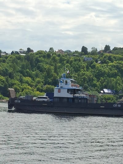 This is a portrait-oriented, landscape photograph taken from a boat or embankment, showing a view of a river and a tree-lined bank with houses on a hill. The dominant subjects are a dark-colored barge with the Cyrillic text "СП-19" visible on its side, and a small white and blue boat with a cabin, moored behind it. The barge appears to be carrying some cargo or equipment, including what looks like a small, brown, box-like structure. The small boat has several windows and a white railing around its deck, with some people or figures visible inside.
The background consists of a dense green forest covering a hillside, dotted with houses. The sky is partially cloudy, with soft white clouds against a pale blue backdrop. The water in the foreground is rippled, reflecting the light and creating a textured surface.
The scene suggests a calm day, likely in the late morning or early afternoon, given the bright but diffused sunlight. There is no indication of any specific activity other than the boats being moored. The location is Verhnij Uslon, Russia, as indicated by the context. The overall mood is serene and peaceful. The visible text "СП-19" on the barge is a prominent detail.