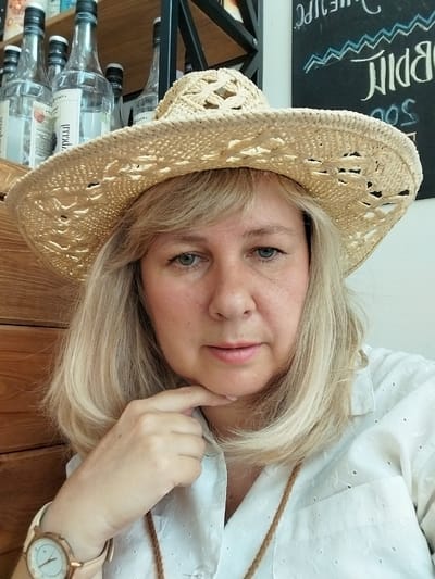 The image shows a close-up, selfie-style shot of a blonde woman wearing a straw hat and a white embroidered shirt. She has her chin resting on her hand, and her gaze is directed towards the camera. In the background, on the left, there are several glass bottles with dark caps arranged on wooden shelves. On the right, a portion of a dark chalkboard with white chalk writing is visible. The lighting is natural, creating some shadows and highlights on the woman's face and the objects in the scene. There is no explicit indication of the location being in the city of Gornyi, Russia.