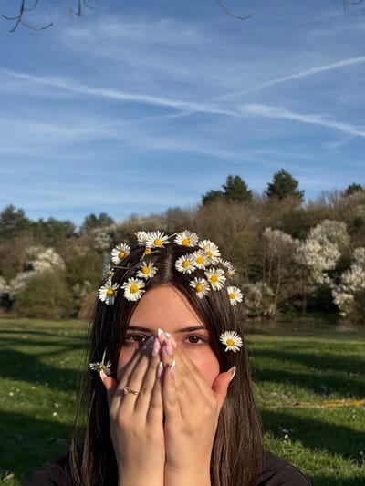A young woman is shown from the chest up. She is outdoors in a grassy field with trees and a blue sky visible in the background. She is wearing daisies in her dark brown hair, arranged like a crown. Her hands are raised to her face, partially covering her mouth. She has an engagement ring on her left hand. The sky is blue with contrails, and the trees are mostly green, though some are bare. The picture was taken in a field with trees in Beli Brod, Bulgaria.