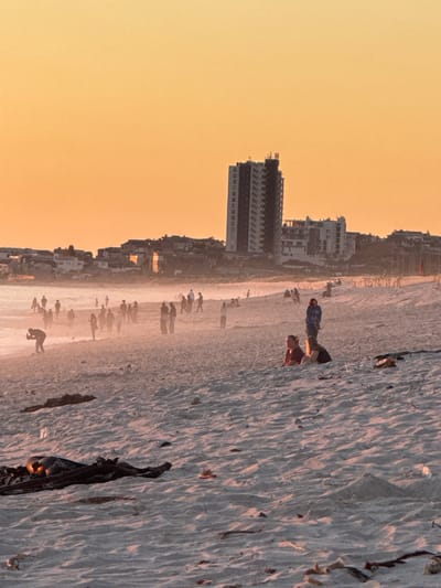 This image depicts a wide, sandy beach bathed in the warm, soft light of the golden hour, suggesting either sunrise or sunset. The sky is a vibrant gradient of yellow and orange, dominating the upper portion of the frame.
Along the horizon line, a dense urban coastline stretches, featuring numerous multi-story buildings that appear to be residential or hotel structures. These buildings are partially silhouetted against the bright sky and the shimmering light reflecting off the beach.
The foreground and mid-ground consist of a broad expanse of light-colored sand, showing subtle patterns and textures. Scattered across the sand are several dark, irregular shapes that could be driftwood or other debris. Numerous people are visible across the beach, from individuals closer to the viewer to smaller, silhouetted figures further in the distance. Some individuals are standing or walking, while others appear to be sitting or looking down at the sand, engaged in leisurely beach activities. For example, two individuals in the mid-ground are clearly visible, one wearing a blue top and another in a striped top, observing something on the sand.
The scene conveys a peaceful and relaxed atmosphere. There is no visible text in the image. The setting is consistent with a bustling coastal area like Blouberg, South Africa, known for its beaches and urban development.