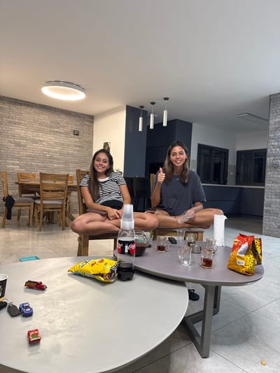 The image displays two young adult females situated indoors at a residence in Yehud Monosson, Israel. Both individuals are seated on chairs with their legs crossed in a lotus-like position, facing the camera. The female on the left wears a black and white striped t-shirt and dark shorts, smiling. The female on the right wears a dark t-shirt, also smiling, and extends her right hand with a thumbs-up gesture.

In the foreground, two nested, elliptical coffee tables are present. The lower table contains a partially consumed Coca-Cola bottle topped with stacked plastic cups, an open yellow bag of cheese-flavored snacks, a set of car keys, and three miniature toy vehicles (a red fire truck, a blue car, and a red/green car). The higher table holds a glass carafe containing a dark liquid, multiple small glass cups (some filled with a dark liquid), a roll of white paper towels, and a red and yellow bag of snacks.

The environment features modern interior design. The flooring is light grey tile. The left wall is constructed of grey brickwork. In the background, a contemporary kitchen with dark cabinetry and dark-framed windows is visible. A wooden dining table and chairs are also partially visible on the left. Illumination is provided by a large, circular ceiling fixture and three dark pendant lights over the kitchen area.
