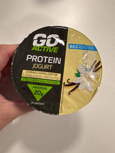 The image shows a close-up, overhead view of a single-serving yogurt container being held by a person's hand. The container has a yellow lid with a stylized drawing of a vanilla flower and bean pods, suggesting a vanilla flavor. Text on the lid indicates "GO ACTIVE PROTEIN JOGURT" and specifies "20g PROTEIN" with "57% ENERGY FROM PROTEIN." Other text, in Polish and potentially other Slavic languages, highlights "BEZ DODATKU CUKRÓW" (without added sugar) and provides a best-before date of "18.07.2026." The overall impression is that of a healthy, protein-rich snack. The background is a plain, neutral surface, likely a table. The lighting appears to be indoor, with no specific time of day or weather cues.
