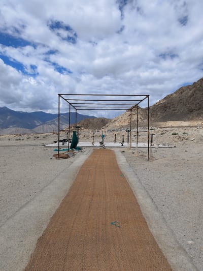 In Phyang, India, an outdoor gym is set up in a barren, mountainous landscape. A sandy path leads toward a metal structure with a thatched roof, possibly an awning or shelter. The gym equipment, made of metal and painted in yellow and green, is arranged on a concrete platform. Mountains form the backdrop, their slopes covered in rocks and sparse vegetation. The sky is filled with scattered clouds, suggesting a partly sunny day. The scene appears to be an isolated area, possibly a rest stop or recreational facility in a remote location.