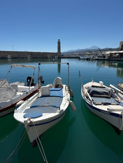 The image shows a view of the Venetian Harbor in Rethymno, Greece. In the foreground, three small boats are docked on the clear, turquoise water. The boat on the left is white with a red stripe along its hull and has an outboard motor. The middle boat is also white, with blue cushions and a canopy. The boat on the right is white with blue trim and has a Greek flag attached to its stern. The middle and right boats are covered with tarps. In the background, a stone breakwater stretches across the harbor, with a lighthouse at its end. Beyond the breakwater, the city of Rethymno can be seen with buildings and mountains in the distance. The sky is clear and blue.