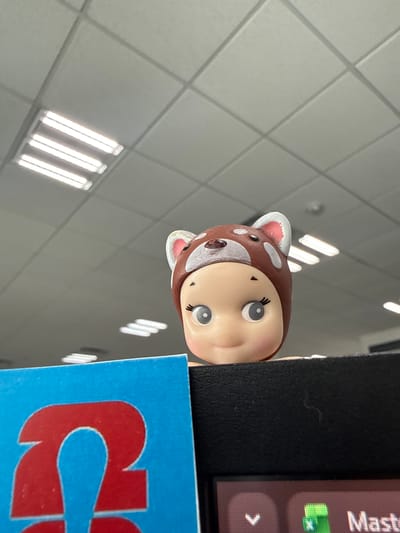The image captures an indoor scene, likely within a commercial or retail establishment, seen from a low perspective looking upwards and to the right. The primary subject is a small, endearing figurine resembling a baby or child dressed in a brown deer or fawn costume. Its head, featuring large, light blue eyes, delicate eyebrows, and a small mouth, is playfully peeking out from behind a dark, textured vertical surface on the right side of the frame. The costume's ears are brown with white spots and pinkish interiors, complementing the white spots on the rest of the brown head covering. The figurine's expression appears gentle and curious.

In the background, a light gray ceiling with a grid pattern of square acoustic tiles is visible. Several rows of bright, illuminated fluorescent light fixtures are embedded in the ceiling, providing strong artificial light to the space.

Adjacent to the figurine, on the right side of the dark vertical surface it is peering over, a digital screen or display is partially visible. On this screen, the upper part of the word "Mast" is visible in white text, along with a green icon and a partial white arrow pointing to the right on a grey background. Below this screen, a vibrant blue surface with a large, partial red letter "C" is seen, likely indicating branding or a sign.

The overall scene suggests a playful arrangement of objects within a brightly lit indoor setting in Sanctorum, Mexico. The image is originally rotated 90 degrees clockwise, giving it an unusual orientation.