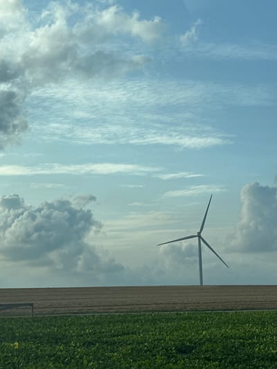 The image captures a wind turbine standing tall in a rural landscape. The scene is divided into distinct horizontal bands. The foreground features a vibrant green field of crops. Above this, a wide strip of brown, tilled earth stretches across the middle ground, likely a cultivated field. The background is dominated by a vast sky filled with a mix of fluffy white and darker gray clouds, interspersed with patches of pale blue. The wind turbine itself is a sleek, modern structure with a tall white tower and three angled blades, positioned in the upper right quadrant of the image, silhouetted against the sky. The overall impression is one of agricultural land and renewable energy generation under a dynamic sky. The image was taken in Saint-Maclou-de-Folleville, France.