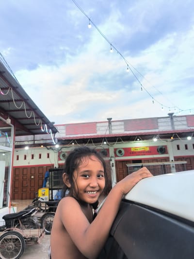 This outdoor photograph, taken in Sukamulia, Indonesia, features a young girl as the central subject. She is positioned in the foreground, looking directly at the camera with a wide, infectious smile that reveals her teeth. Her dark hair appears wet or damp, and her shoulders are bare. She is leaning on the white roof and black trim of a vehicle, which partially fills the lower right section of the frame.

The background depicts a semi-urban scene with a series of buildings, possibly shops or houses, under a cloudy sky. These structures feature red-tiled roofs, white walls, and dark brown doors with small, square windows. Some buildings are adorned with decorative, colorful strip lights. A prominent red sign on one building reads "REX TEUWO," "CAFE ESPRESSO," and "CERIAM." Overhead, strings of illuminated bare light bulbs stretch across the frame, suggesting the time of day is late afternoon or early evening. To the left, a motorcycle with a sidecar or modified cargo attachment is visible. The overall mood is cheerful, accentuated by the girl's happy expression amidst a lively local setting.