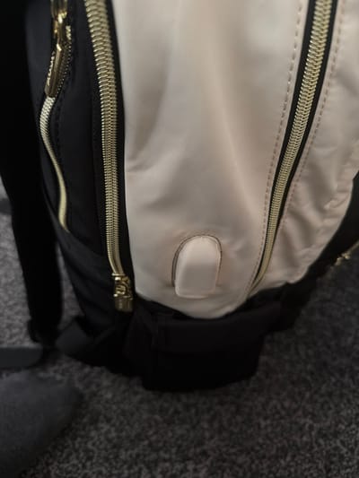 The image shows a close-up view of a partially open black and beige backpack lying on a gray carpet. The zippers are gold-colored. The beige portion of the backpack has a small rectangular patch sewn onto it. The background is a textured gray carpet. There are no people visible in the image. The location is not discernible from the image.