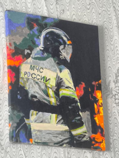 The image is a painting depicting a firefighter in a helmet and uniform with reflective strips. The uniform has a yellow rectangular patch with Cyrillic text that reads "МЧС РОССИИ" which translates to "Russian Ministry of Emergency Situations". The background is abstract with dark and fiery colors suggesting a scene of emergency or fire. The painting is placed against a textured white background, possibly a wall or fabric. There is no discernible location in the city of Psebai, Russia depicted in the artwork itself.