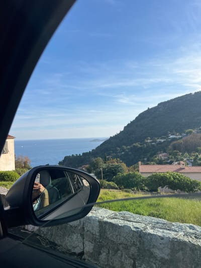 This image captures a scenic coastal landscape, viewed from the interior of a car, likely along a hillside road in Èze, France. The primary vista features a vast expanse of deep blue sea stretching towards the horizon under a bright blue sky adorned with wispy white clouds. On the left, a steep, tree-covered mountain slopes down, dotted with houses that have distinctive terracotta tiled roofs.

In the immediate foreground, the dark frame of the car's window and dashboard is visible in the lower left. The car's side mirror is prominent on the right, offering a reflection of both the car's interior and the surrounding environment. Within the mirror, an arm and hand, possibly wearing a bracelet, are visible holding a smartphone that appears to be an iPhone with a gold-colored ring or case. The reflection also shows a glimpse of the car's seat and a grey, textured stone wall or rock formation by the roadside, which is also visible directly outside the car on the right, partially covered by grass and sparse vegetation.

The weather is clear and sunny, indicative of daytime. The overall impression is one of a leisurely drive through a beautiful Mediterranean setting. No legible text is visible within the image.