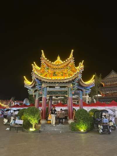 At night in Eshan Yi Autonomous County, China, a brightly lit, ornate pavilion with a multi-tiered roof and intricate details stands as the central focus. Yellow lights outline the roof's curves and decorative elements. Two individuals, a woman in a black t-shirt and cream-colored pants carrying a yellow bag, and a person in a brown jacket and black pants, are walking on the steps of the pavilion. Surrounding the pavilion are lush green bushes, some illuminated by spotlights. To the left of the pavilion, a bench sits near a white electric tricycle, and further back, red tents and more vehicles suggest a marketplace or event. To the right of the pavilion, another electric tricycle is parked. In the background, traditional Chinese architecture with illuminated roofs is visible. The ground is paved, and the overall atmosphere is festive and illuminated.
