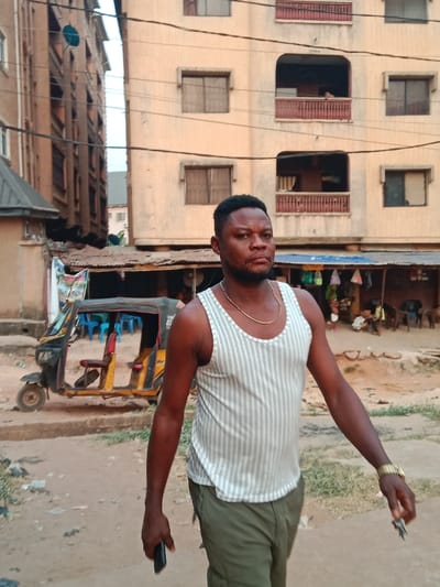 A man with short black hair, wearing a striped tank top and khaki pants, walks on a dirt road. He has a gold chain and watch. Buildings stand behind him, one with multiple windows and balconies. A three-wheeled vehicle is parked nearby. A covered market area is visible in the background. The man holds a phone in one hand and keys in the other.