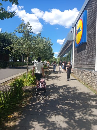 In Laxou, France, on a sunny day, a group of people are walking along a sidewalk in front of a Lidl supermarket. The sun casts dappled shadows of tree leaves onto the gray concrete path.
The building's facade is a combination of wooden panels and a section with a stone-like material. Above the entrance, a prominent, circular blue and yellow Lidl logo is visible. Several people are walking, some appearing to be a family or friends. One individual in the foreground is pushing a shopping cart with a patterned bag. Another person in the background is walking towards the supermarket. The sky is bright blue with fluffy white clouds, indicating good weather. The overall scene suggests a typical daytime shopping excursion. The time of day appears to be late morning or early afternoon, given the length and angle of the shadows.
