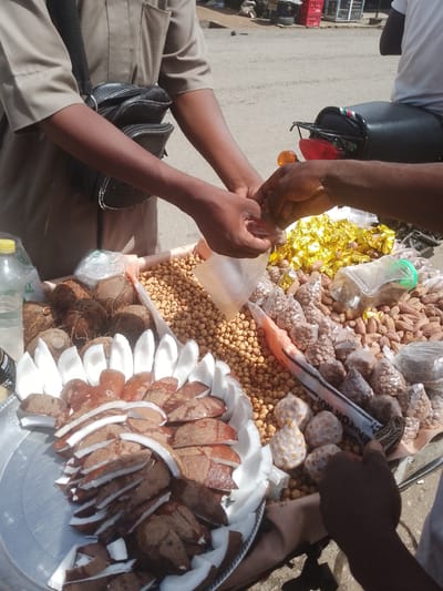 This image depicts a scene of commerce in Beli, Nigeria. The foreground is dominated by a display of food items, primarily coconuts cut into slices and arranged on a metal platter. Several whole coconuts are also visible, alongside small bags of nuts or seeds and a large pile of what appears to be roasted chickpeas. There are also shiny, gold-wrapped candies. The items are laid out on a surface, likely a cart or stall, with a portion of a motorcycle seat visible in the background, suggesting an outdoor market setting.
Two individuals are engaged in a transaction. One person, wearing a light brown uniform and a black bag slung across their chest, is handing items to another person whose hands are partially visible. The interaction appears to be a purchase, with the seller transferring goods to the customer. The lighting suggests it is daytime, with bright sunlight casting shadows. The overall atmosphere seems busy and functional, typical of a market environment. No visible text is present in the image.