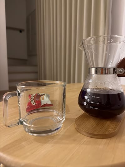 This is a close-up, eye-level, indoor photograph of a wooden table with two coffee-related items. On the left is a clear glass mug containing a single, small, individually wrapped chocolate bar. The mug has a handle. On the right is a glass pour-over coffee maker filled with dark coffee, resting on a light-colored wooden base. The coffee maker has measurement markings in milliliters, with the coffee level appearing to be around 350 ml. The background features a cream-colored, vertically pleated curtain and a dimly lit staircase with a handrail. The location is not specified in the image or the provided context of Bang Kaeo Subdistrict, Thailand.
