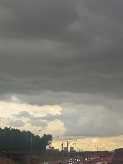 This media file captures a dramatic sky over a highway in Novodrozhzhino, Russia. The upper portion of the image is dominated by heavy, dark gray storm clouds, suggesting an impending downpour. Below these ominous clouds, a lighter band of sky appears, illuminated by what seems to be the fading light of late afternoon or early evening, possibly with some breaks allowing sunlight through.
The scene is set on a multi-lane highway, with a visible overpass or bridge spanning across it. Along the highway, there is significant traffic, indicated by the rows of illuminated vehicle lights, suggesting congestion or a busy commute. Numerous streetlights, mounted on poles, line the highway, casting a warm glow that contrasts with the cool tones of the sky.
A dense line of dark green trees borders the highway and the overpass, providing a natural element against the man-made structures and the dramatic weather. The overall mood is somewhat somber and dramatic, largely due to the imposing storm clouds. The time of day appears to be either late afternoon or early evening, given the quality of light. There is no visible text in the image.