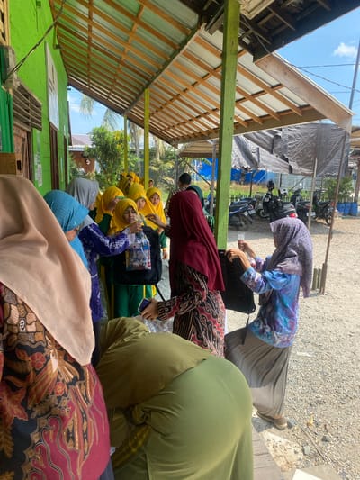 The image depicts an outdoor scene likely in Banyu Hirang, Indonesia, beneath a covered structure. Several individuals, predominantly women and girls, are gathered. Many are wearing hijabs and traditional Indonesian attire, such as batik.
In the foreground, on the left, a person wearing a beige hijab and a batik top is partially visible. To their right, a woman in a blue and white patterned top and a grey pleated skirt is interacting with a black backpack. She is holding a clear plastic bag containing items.
A group of younger individuals, mostly wearing yellow hijabs and green school uniforms, are in the middle ground. One girl in a yellow hijab is reaching for or receiving a clear plastic bag from someone out of frame. Another young girl is looking at the bag.
In the background, several motorcycles are parked. The ground is covered with gravel. A green building is visible on the far left, with wooden beams and corrugated metal roofing forming the covered area. The sky is clear and blue with some clouds. The overall impression is of a community gathering or distribution event.