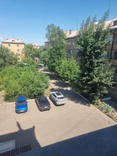 The image shows a residential courtyard in the city of Saran, Kazakhstan. Several cars are parked in the courtyard, including a blue sedan, a dark sedan, and a white SUV. The buildings are typical of Soviet-era apartment blocks, with yellow brick facades and pitched roofs. There are many trees and bushes, suggesting a green and well-maintained environment. The sky is clear and blue, indicating a sunny day. In the foreground, a shadow of a person is visible on the pavement, indicating that someone is likely observing the scene from a higher vantage point, possibly a balcony or window.