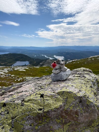 A cairn of stacked rocks, topped with a splash of red paint, rests on a lichen-covered boulder in the foreground. The cairn marks a scenic overlook in Ormemyr, Norway, with a vast landscape of rolling hills, forests, and lakes stretching into the distance under a partly cloudy blue sky. The terrain is rugged, with patches of green vegetation and scattered rocks. The atmosphere is serene and natural.
