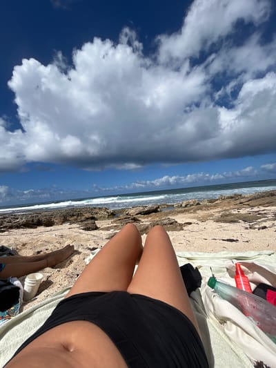 The image depicts an outdoor beach scene in Le Moule, France, viewed from a low-angle, first-person perspective. In the immediate foreground, the lower torso and upper thighs of an individual with tan skin are visible, clad in black swim bottoms. This individual is lying on a light-colored towel. To the right on the towel, a clear plastic bottle with a green cap, appearing partially filled with liquid, and a red spray bottle are present. To the left, further into the midground, the lower legs and feet of a second individual are visible, resting on the sand, with a thin bracelet on one ankle. Adjacent to this person's feet, a white disposable cup and a multi-colored cloth are on the sand. The beach consists of light beige sand interspersed with small shells and dark reddish-brown organic debris. Beyond the sandy area, a rugged, dark brown rocky shoreline transitions into the ocean. The ocean features blue-green water with multiple lines of white-capped waves breaking towards the shore. The sky above is bright blue, occupied by large, fluffy white cumulus clouds, with some areas exhibiting darker grey cloud formations.
