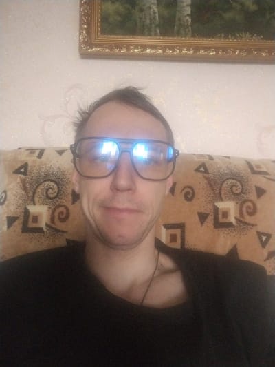 The image depicts a close-up, front-facing portrait of an adult male in an indoor residential setting, purportedly in Furmanov, Russia. The subject has short, dark hair and light skin. He is wearing large, dark-framed aviator-style glasses, exhibiting prominent blue-white light reflections that obscure his eyes. He wears a dark, possibly black, crew-neck shirt, with a thin, dark necklace visible at his collarbone. The man exhibits a slight, closed-mouth smile. He is seated or leaning against a piece of furniture, likely a couch or armchair, upholstered in a brown-toned fabric with a repeating pattern of dark geometric shapes, including squares, triangles, and swirling motifs. Behind him, a light-colored wall is visible, adorned with a subtle, slightly reflective patterned wallpaper featuring faint scrolls or floral designs. Above and to the right of the subject's head, a decorative gold-framed picture hangs on the wall, depicting a natural landscape with several light-barked trees, consistent with birches.