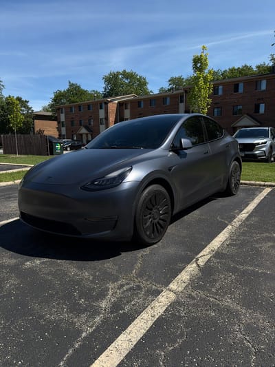 A matte gray Tesla is parked in an asphalt parking lot with white painted lines. The car is angled towards the right of the frame, with its front left wheel visible and a portion of the rear wheel. The car has dark tinted windows and black rims. In the background, there is a red brick apartment building with multiple windows. Trees with green foliage are visible between the car and the building, as well as in front of the building. The sky is clear and blue. To the left of the Tesla, another silver car is parked. Further to the left, a green dumpster with the "GROUT DOCTOR" logo is visible. The scene appears to be set on a sunny day, likely during daylight hours. The location is Vernon Hills, USA. There are no people visible in the image.