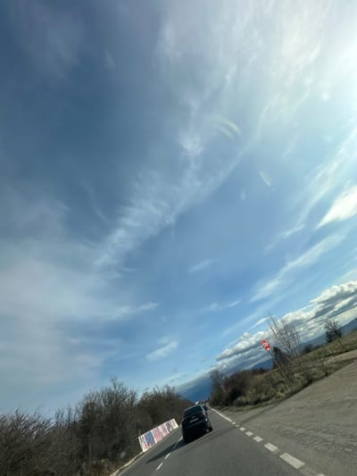 A wide-angle, low-angle photograph captures a rural road scene under a partly cloudy sky near Bogyovtsi, Bulgaria. The asphalt road extends into the distance, with a dark-colored hatchback automobile traveling away from the camera in the right lane. The road features a solid white line on the right shoulder and dashed white lane markers. To the left of the road, dense, bare deciduous vegetation forms a treeline, behind which a white and striped barrier or fence is partially visible. To the right of the road, sparse bare trees and shrubs are present, followed by open, grassy terrain that slopes upwards towards distant hills or mountains on the horizon. A red octagonal "STOP" sign on a white pole is positioned on the right shoulder. The sky occupies the majority of the frame, displaying various formations of wispy white clouds against a bright blue background, with a strong light source, likely the sun, emanating from the upper right quadrant. No individuals are visible.