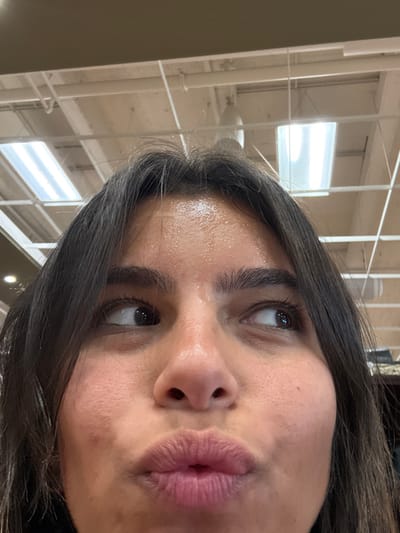 A close-up, low-angle photograph captures a female individual from the forehead to the upper lip, with dark hair framing the face. The forehead exhibits a shiny appearance, suggesting perspiration. Both eyes are looking upward and slightly outward, with the left eye directed towards the upper left and the right eye towards the upper right. The lips are pursed, forming a "kissing" or "fish face" expression. The visible background is an interior ceiling featuring exposed white pipes and multiple rectangular fluorescent light fixtures, characteristic of a commercial or institutional building. The scene is located in Plainsboro Township, United States.