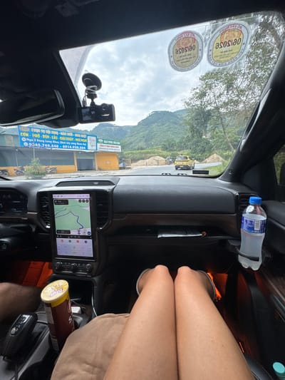 Captured from inside a vehicle, the image shows a view through the windshield, presenting a mountainous landscape under a cloudy sky. Visible through the front window is a road with a yellow vehicle and construction materials. The dashboard features a navigation screen displaying a map. In the foreground, parts of a person's legs are visible. Other items include a bottle of water and a container of Lay's Stax. A promotional banner is seen on the side of the road, which mentions services like "COM - PHO - BUN - LAU - GIAI KHAT - NGU NGHI", and the phone numbers "0345.749.540 - 0914.920.504" as well as the location "Ngà 3 Đông Lùng Khuy (Qua cây xăng Quản Bạ)", which indicates that this is the Minh Tân Commune in Vietnam.