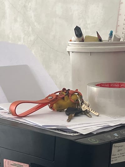 The image depicts a close-up of a desk surface with various office supplies and personal items. In the foreground, a set of keys hangs from a yellow keychain attached to an orange strap, resting on a stack of white papers. To the right of the keys, a roll of clear tape with red branding is visible. Behind the keys and tape, a white plastic bucket holds several writing utensils, including a black marker, a pencil with a pointed tip, and a blue pen. A ruler is partially visible on the far right. The background consists of a textured, light-colored wall. The overall scene appears to be a personal workspace. There is no specific indication of a location in Boja, Indonesia.