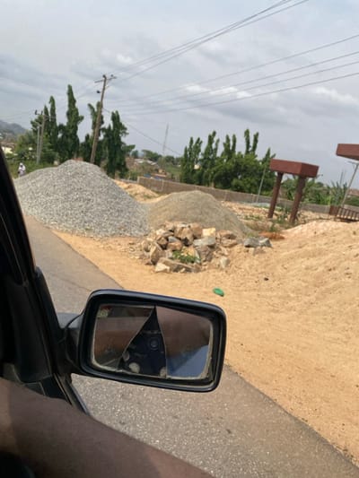 The image depicts an outdoor scene in Okengwe - Obehira, Nigeria, captured from the passenger side of a dark-colored vehicle. A dark-skinned arm is visible in the lower-left. A rectangular side-view mirror with significantly fractured glass, broken into multiple distinct pieces, is centrally positioned in the foreground. Outside the vehicle, a gray paved road transitions to a sandy, undeveloped terrain. This terrain features a large conical mound of light-gray gravel, a smaller mound of finer, lighter-colored aggregate, and a collection of rough-hewn rocks. A small green plastic item rests on the dirt ground near the center-right. In the background, tall green trees, multiple utility poles supporting several overhead power lines, and distant hills with scattered structures are visible. Two brown, rectangular structures on angled supports are partially seen in the far-right background, suggesting elements of a construction project. A lone individual, dressed in white attire, is faintly discernible walking on a distant path near the leftmost gravel pile. The sky is uniformly overcast.