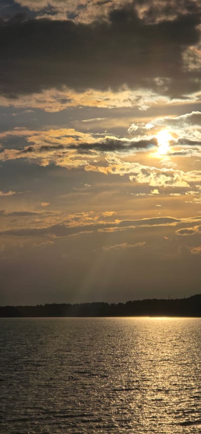 The image depicts a serene lake at sunset, with a dark, silhouetted treeline forming the horizon. The sky is filled with dramatic, textured clouds, illuminated by the golden rays of the setting sun which create a shimmering path of light across the water. The scene is devoid of any human presence or activity. The location is identified as Pristan'-Przheval'sk, Kyrgyzstan.