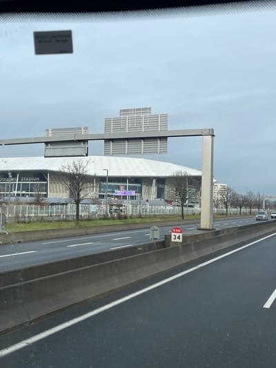 The image, captured from the interior of a vehicle, depicts a section of the N346 highway in Décines-Charpieu, France, under an overcast sky. The foreground shows multiple asphalt road lanes with white painted markings, bordered by a concrete median barrier. A red and white marker sign on the median indicates "N346 34". An overhead grey metal gantry spans the highway, supporting several blank rectangular sign panels.
To the left of the highway, beyond a grassy verge and a fence, stands the Groupama Stadium. This is a large, modern multi-level stadium with a white, slightly domed roof and a façade composed of grey structural elements and large glass sections. The name "Groupama STADIUM" is prominently displayed on the building. Near the stadium's apparent entrance, digital signs show "P1 P2 P3". Several bare deciduous trees are positioned between the stadium complex and the highway.
On the right side of the image, beyond the median, parallel highway lanes extend into the distance, occupied by a few white and grey civilian vehicles. Further right, a row of bare trees and a multi-story building are visible in the background. A small, partially obscured rectangular sticker on the upper interior of the vehicle's windshield displays numerical values associated with "KM/H" and "MPH". No individuals are discernible within the scene.