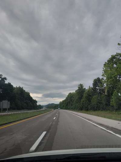 The image depicts a highway scene in Clay City, United States of America, under a cloudy sky. The perspective is from inside a vehicle, looking forward along a two-lane highway. Trees line both sides of the road, with dense green foliage. In the distance, rolling hills are visible under the overcast sky. Several vehicles are present on the highway, including a white van and a black SUV, traveling in the same direction as the viewer. The road surface shows some signs of wear and moisture. A yellow line delineates the shoulder on the left side of the road, and white lines mark the lanes. A signpost is visible on the left shoulder.