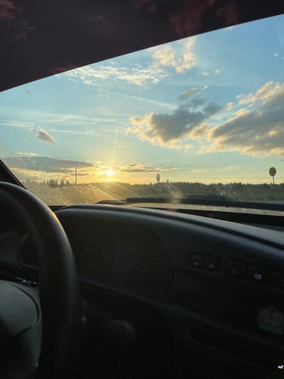 The image depicts a view from inside a vehicle, looking out through the windshield. The sun is setting, casting a warm glow across the sky, which is filled with scattered clouds. The scene outside appears to be a rural or semi-rural landscape with trees and some utility poles visible in the distance. There are also two circular signs visible along the roadside. Inside the car, the steering wheel and part of the dashboard are visible, indicating the perspective of the driver or a passenger in the front seat. The image is taken in the city of Sovkhozny, Russia.