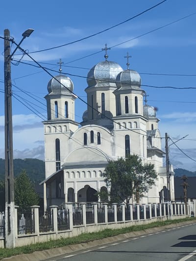 A white Orthodox church with multiple domes topped with crosses stands under a bright blue sky with scattered clouds. The church has several towers and arched windows. A black metal fence with white pillars encloses the church grounds, with a sidewalk and a road in the foreground. Power lines crisscross the sky in front of the church. The church is located in Petrova, Romania.
