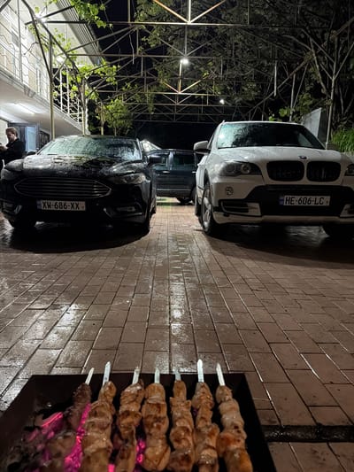 The image shows an outdoor scene at night, likely in Kapreshumi, Georgia, based on the vehicle license plates. Two cars, one white and one black, are parked on a brick or stone surface that appears wet, possibly from rain. In the foreground, skewers of meat are being grilled over hot coals in a barbecue. The skewers are placed close to the grill's edge. Above and to the left of the black car, there's a structure with climbing plants. A person is partially visible near the black car. The license plate on the white car reads "HE-606-LC" and the black car's license plate reads "XH-686-XX". The overall lighting is dim, illuminated by nearby lights and the glow from the barbecue.