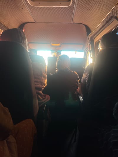 The image shows the interior of a vehicle, likely a van or bus, seen from the perspective of a passenger. The ceiling of the vehicle is covered in a diamond-patterned upholstery, and there is a sun visor with a yellow light fixture. Sunlight streams in through the front windshield, creating lens flare and silhouetting the passengers.

Several passengers are visible, mostly from behind. In the foreground, a dark-colored seat is partially visible on the left. Further back, a person wearing a hooded garment is seated in the center, their back to the camera. To their right, another passenger is wearing a white t-shirt. On the far right, another silhouetted passenger is seated. A person in the middle ground on the left appears to be wrapped in a patterned blanket. The overall impression is of a group of people traveling together, possibly on a journey through or near the city of Nuro, Kyrgyzstan, given the prompt's reference. The lighting suggests it is either early morning or late afternoon.