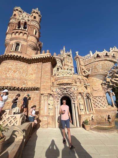 The image depicts Colomares Castle, also known as Monumento a Colón, in Benalmádena, Spain. The castle is a monument dedicated to Christopher Columbus. In the foreground, a young man wearing a pink sleeveless shirt and blue shorts stands looking at his phone. He is wearing sunglasses and black sandals. Several other individuals are visible in the background: a woman with a white hat and white shorts on the far left, a woman in a black tank top and jeans next to her, a person in a dark t-shirt and black shorts, and a woman in a white tank top and denim shorts. They appear to be tourists exploring the castle grounds. The architecture of the castle is intricate and detailed, constructed from stone and brick, with various turrets, arches, and decorative elements. The sky is clear and blue. The scene is set during daylight hours.