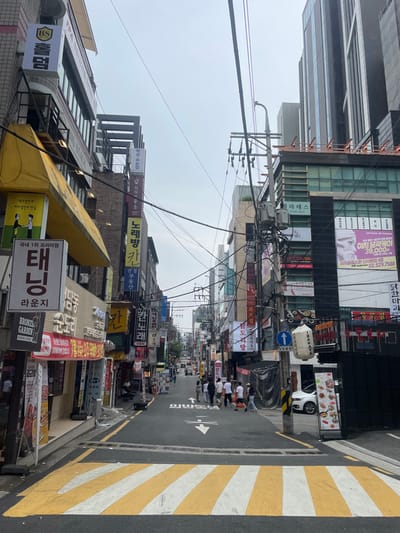 The image displays an urban street scene in 강남구 (Gangnam-gu), South Korea, under an overcast sky. The perspective looks down a narrow, ascending asphalt street lined densely with multi-story commercial buildings. In the immediate foreground, a yellow and white striped crosswalk extends across the road.
On the left, a building features a prominent yellow awning, below which a large white vertical sign displays "태닝" (Tanning), "라운지" (Lounge), and "BRONZE GARDEN". Additional signs and a poster display are visible on the building and sidewalk. On the right, a utility pole is heavily instrumented with multiple transformers and a dense network of overhead power and communication lines. A blue circular traffic sign indicates straight-ahead and right-turn directions. A white cylindrical lantern, marked "INAZAYA", hangs from a structure. A white SUV is parked partially visible on the right, alongside a freestanding menu board.
In the midground, a group of approximately ten pedestrians, primarily young adults, walks collectively on the right side of the street, moving towards the foreground. One individual is positioned slightly ahead and to the left of this group. The buildings lining the street display numerous Korean business signs for establishments such as "노래방" (Noraebang/Karaoke), "부동산" (Real Estate), and various restaurants and beauty services. Further down the street, additional pedestrians and vehicles are discernible. The street continues into the background, flanked by more commercial structures, with taller, modern buildings visible against the skyline in the distance on the right.
