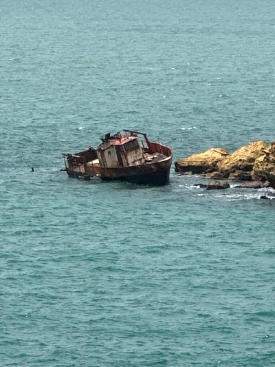 A partially submerged, rusted shipwreck rests in choppy, teal-colored water. The vessel, appearing to be a small fishing boat or similar craft, is tilted to the port side, with its superstructure heavily damaged and appearing derelict. The hull is dark and appears to be made of metal, showing significant corrosion. To the right of the shipwreck, a cluster of rough, tan-colored rocks juts out of the water, with smaller rocks and waves breaking around them. The sea is a uniform teal color with whitecaps indicating moderate wind. In the distance, the water appears calmer. There are no visible people in the image. The scene is desolate and suggests abandonment and decay. The location is not discernible from the image itself and therefore cannot be placed in Uribia, Colombia.