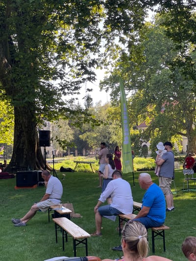 The image depicts an outdoor event in Stupava, Slovakia, likely a park or public garden, with a large tree dominating the left side of the frame. Several people are present, some seated on wooden benches, others standing. In the foreground, a man in a white shirt and beige shorts sits on a bench, looking to his right. Beside him, another man in a blue t-shirt and dark shorts is seated on a separate bench. In the middle ground, a group of people stand near a tall, green banner. A man in a grey t-shirt holds a baby in a white sun hat. Further back, a stage setup with speakers and equipment is visible near the base of the large tree. The background shows more trees, buildings, and a general park-like setting. The lighting suggests it is a sunny day.