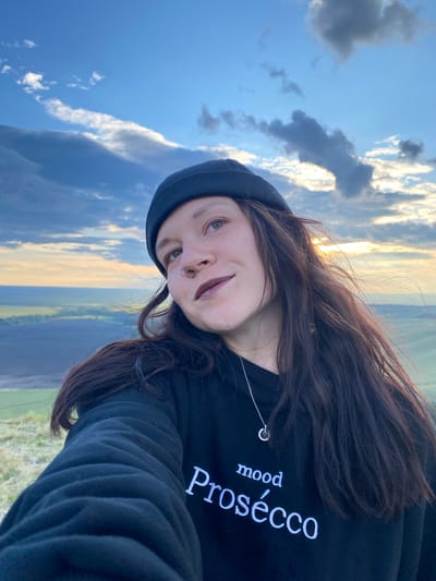 A young person wearing a black beanie and a black hoodie with white text that reads "mood Prosecco" is captured in a selfie from the chest up. They are looking up and to the right, with their head tilted slightly. The background is a vast expanse of sky filled with dramatic clouds, with a hint of sunset glow peeking through. Below the sky, a landscape of rolling fields can be seen, suggesting an outdoor, possibly rural setting. The location is specified as Абзаново, Russia.