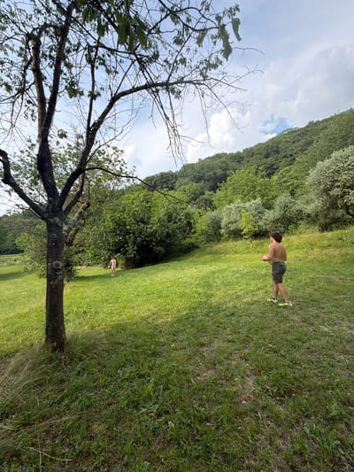A shirtless person stands on a grassy slope, holding a pink object in their right hand. They are wearing dark shorts and bright yellow sneakers. In the background, another person is visible further up the slope, also on the grass. The scene is set in a lush, green environment with trees and vegetation covering the rolling hills. The sky is partly cloudy with a mix of blue and white. The location is believed to be Abbadia Lariana, Italy.